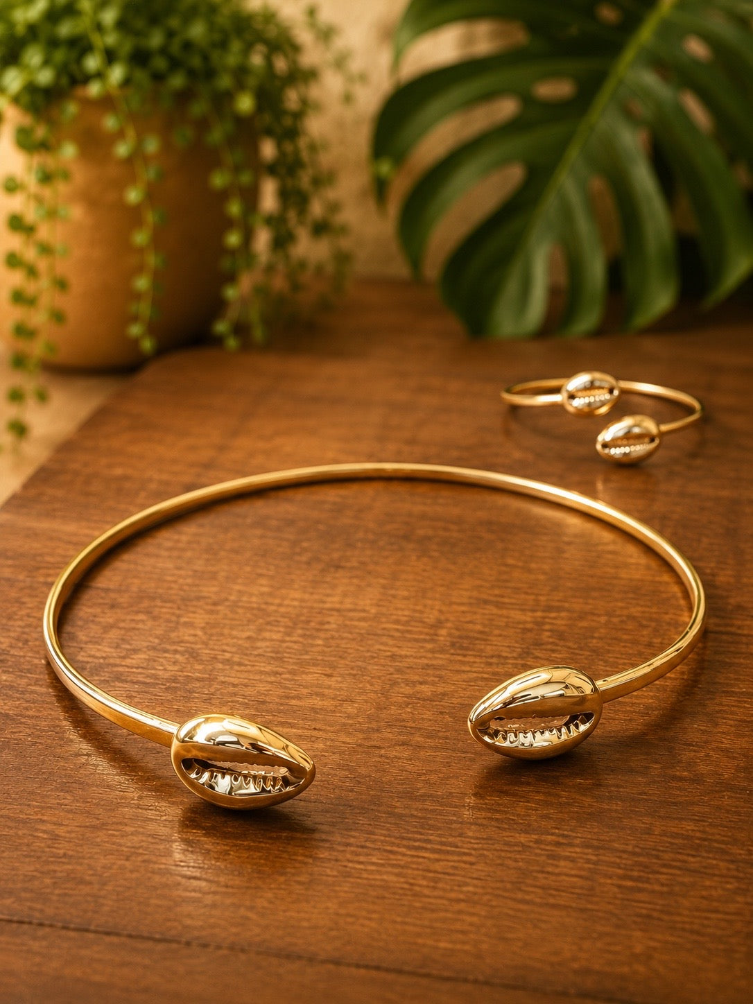 brass cowrie shell choker necklace with brass shell bracelet designs on a wooden surface with plants in the background