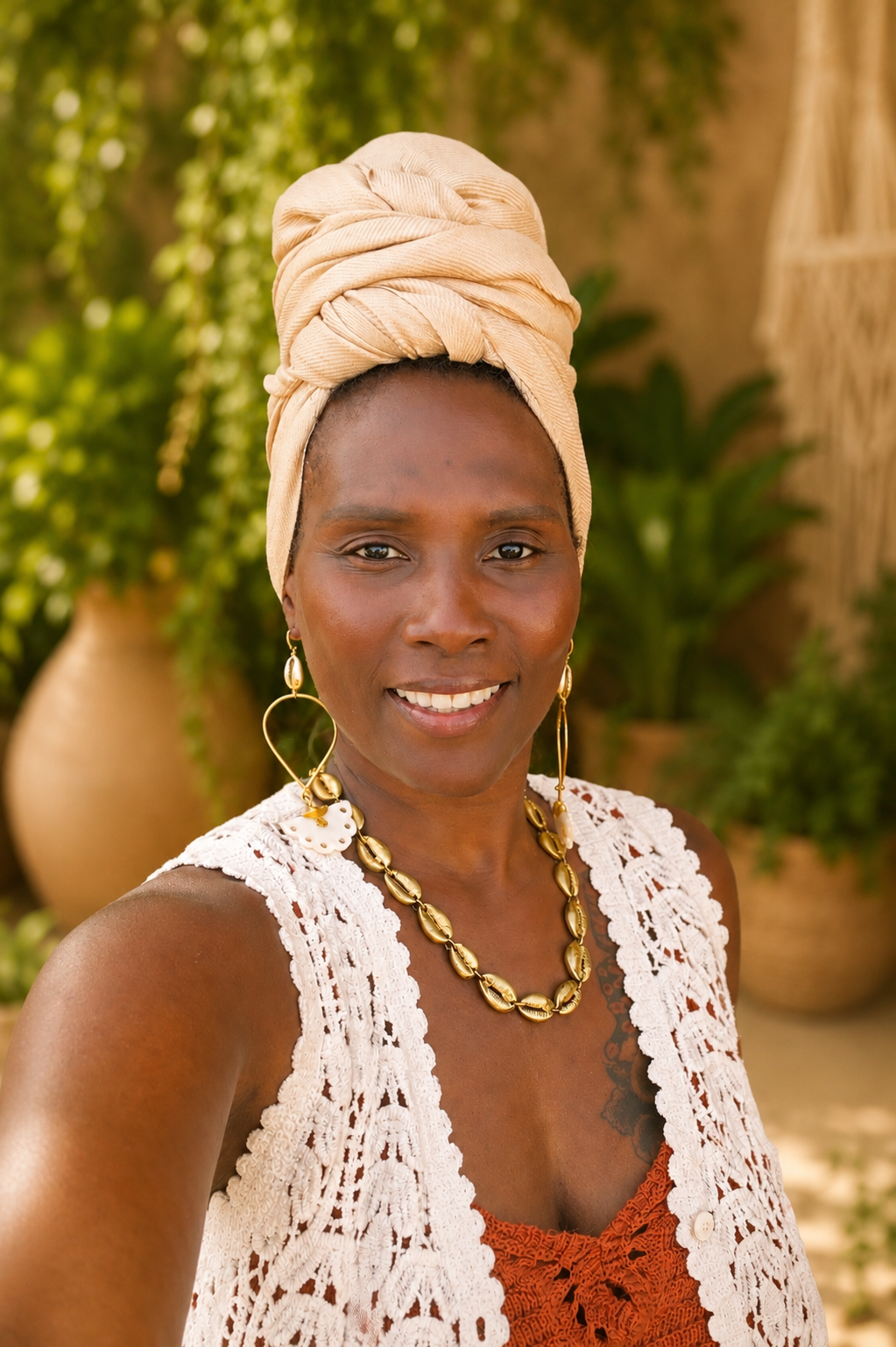 Woman wearing a beige headscarf and brass jewelry outdoors with plants in the background