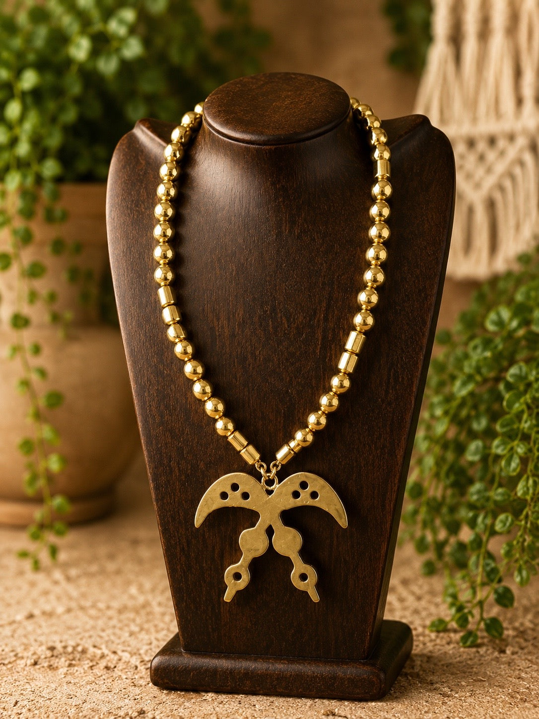 brass necklace with a pendant on a wooden stand with plants in the background