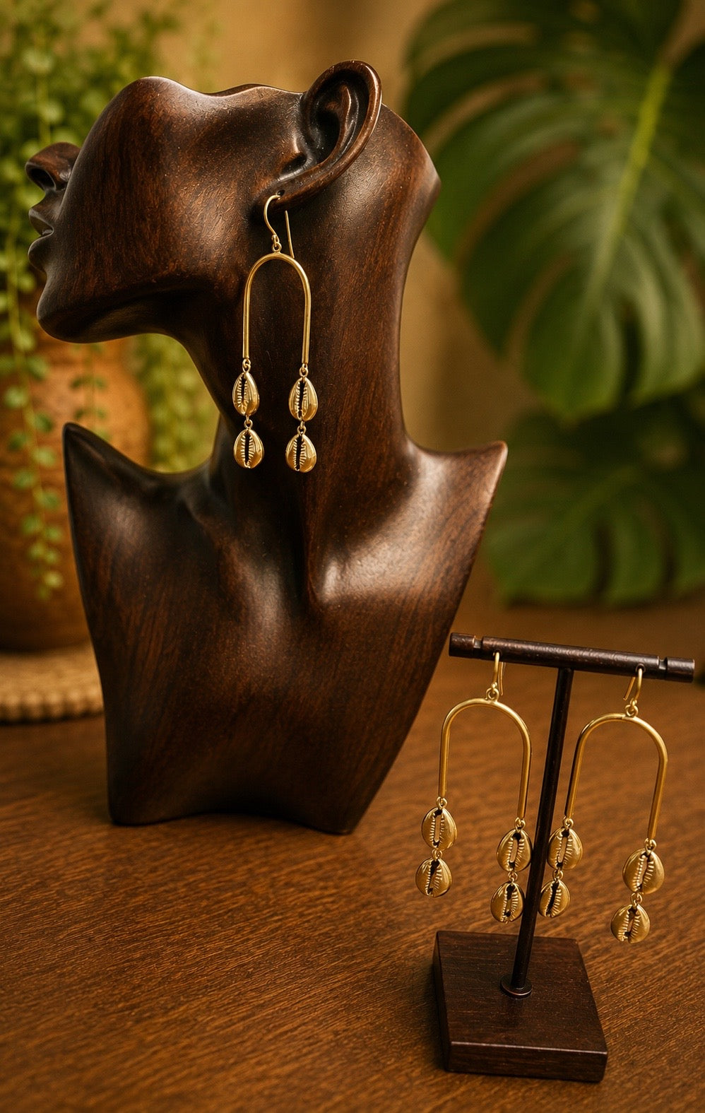 brass cowrie shell earrings on a wooden bust and stand with a blurred plant background